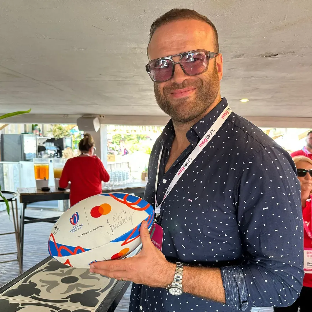 Man holding a decorated cake at an outdoor event, wearing sunglasses and a dark patterned shirt.