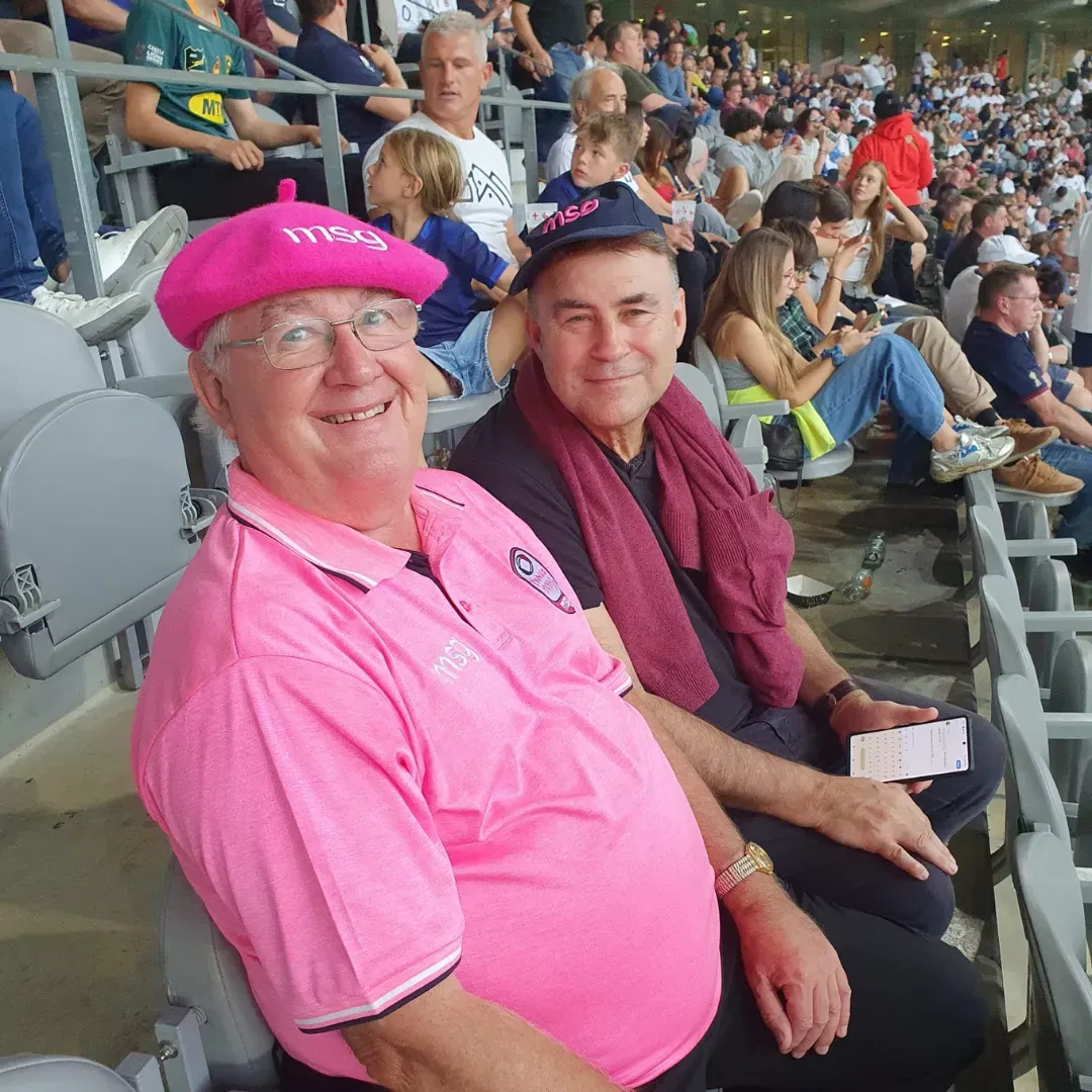 Two spectators in pink and maroon sitting in stadium seats, smiling at a sports event