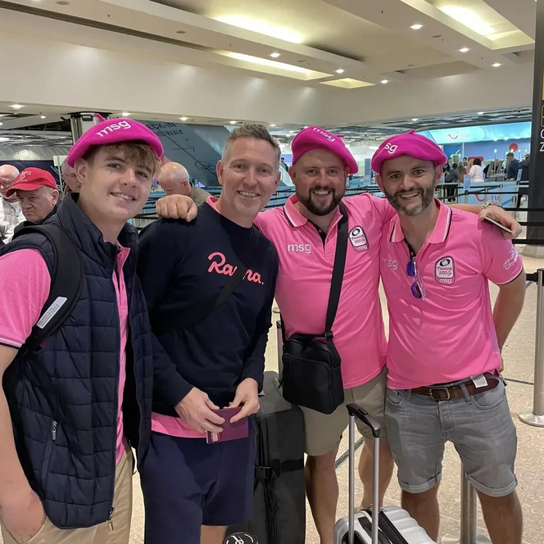 Four smiling friends in pink shirts and caps posing indoors at a lively event.