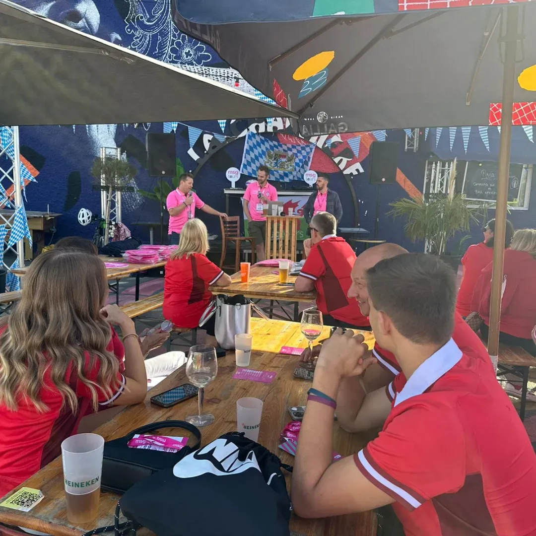 People seated at outdoor tables watching a pink-shirted band perform on a small stage under umbrellas.