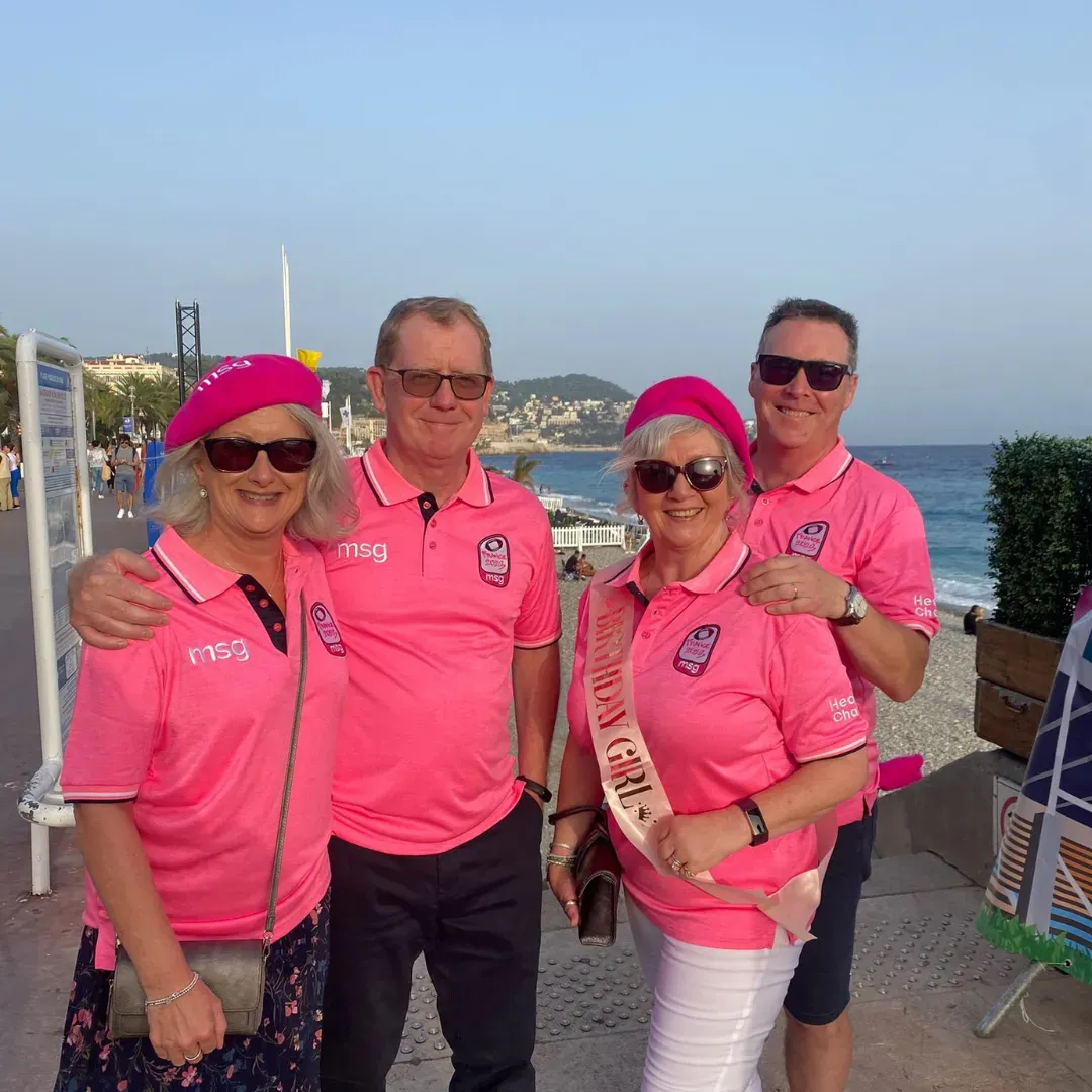 Four people in bright pink shirts posing outdoors by the sea, smiling in sunglasses and hats.