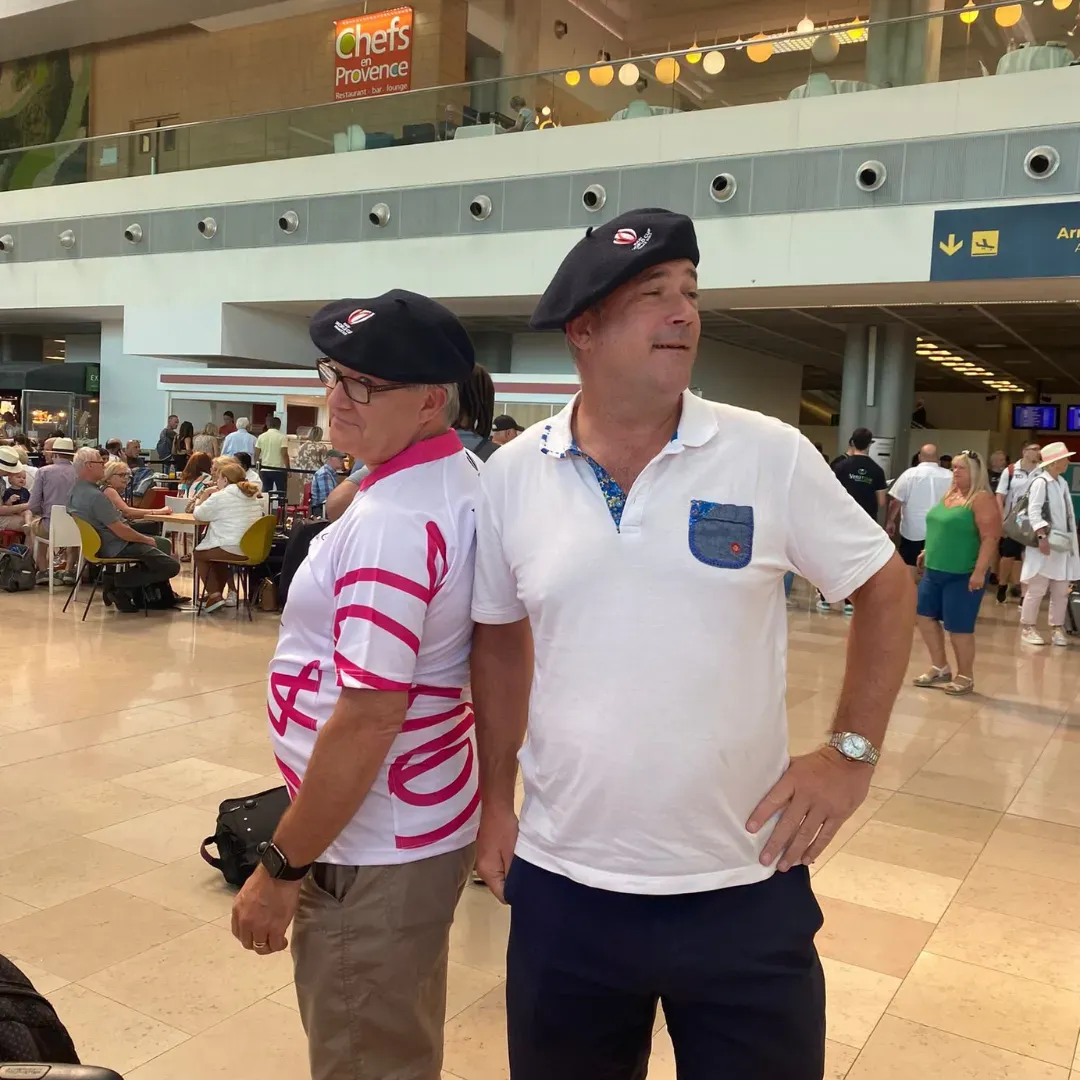 Two men in caps standing in a busy indoor airport terminal, one in a white polo and one in a striped shirt.