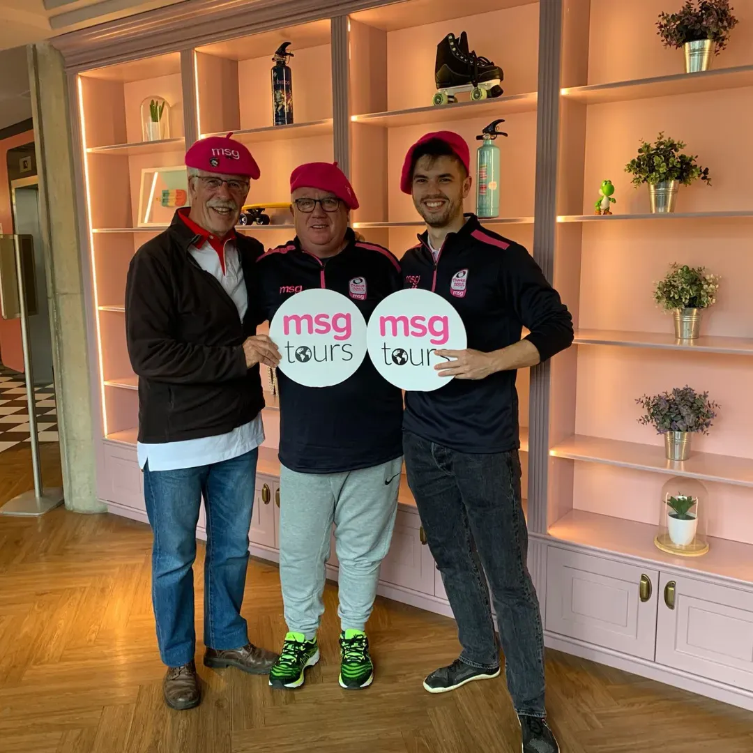Three people in pink hats hold “msg tour” signs in a warmly lit room with shelves and potted plants.