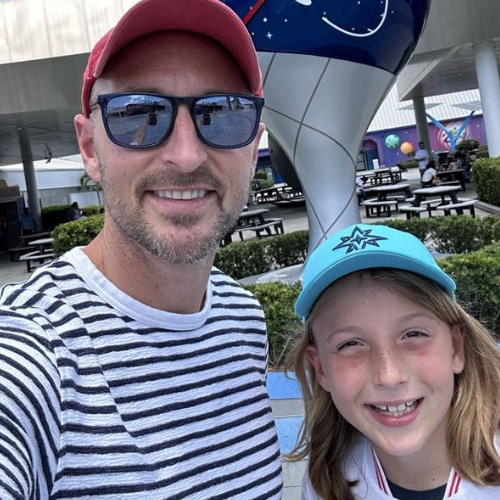 Man and girl smiling in sunglasses and caps at an outdoor amusement park.