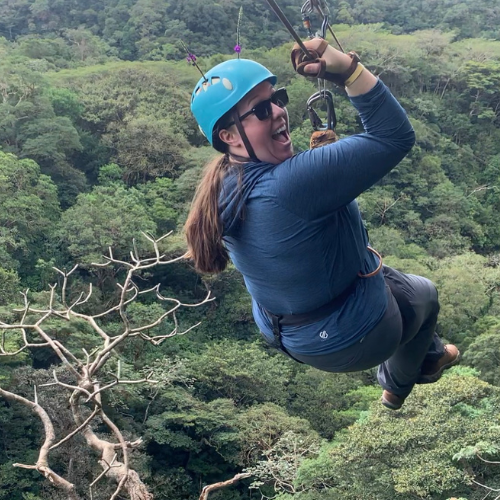 Person ziplining over a lush green forest, wearing a blue helmet and jacket, smiling midair