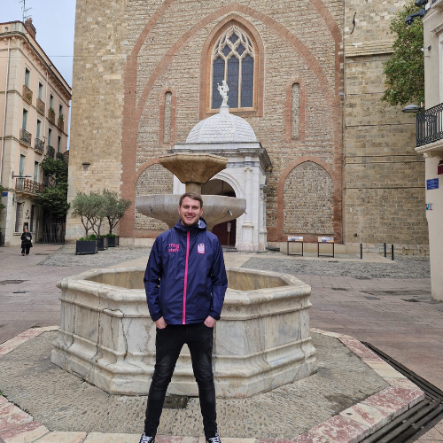 Person standing by a stone fountain in a plaza in front of an old brick church, wearing a navy jacket.