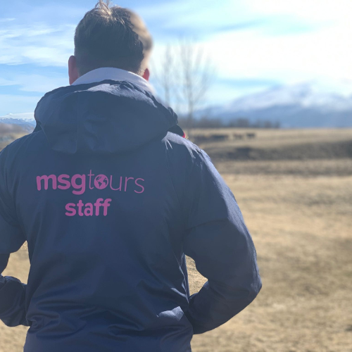 Person in a navy jacket with “msg tours staff” on the back, standing outdoors in a field with mountains.