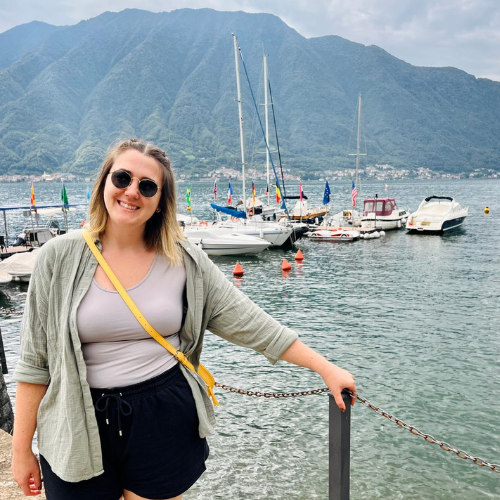 Smiling woman in sunglasses by a marina, with boats and mountains in the background