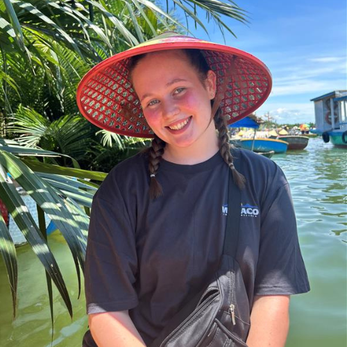 Smiling person in a red sunhat by tropical water with palm leaves and boats in the background