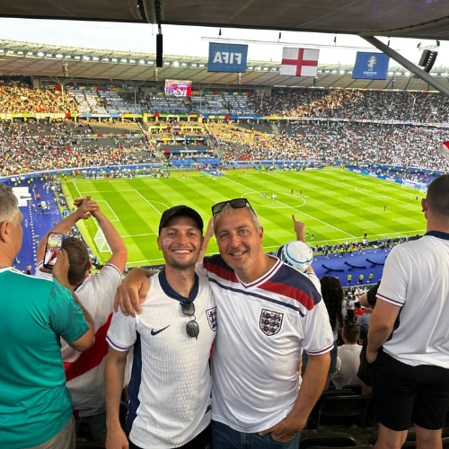 Two fans in white England jerseys smiling at a packed soccer stadium, one raising a drink.