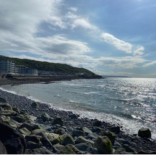 Rocky seaside shore with waves, cloudy sky, and buildings along a green hillside