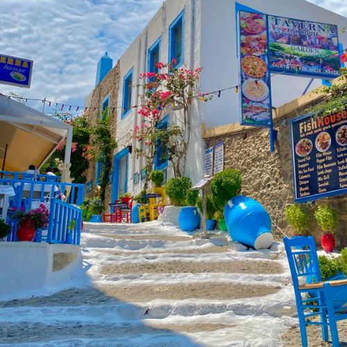 Bright white steps lead to a blue seaside restaurant with colorful signs and potted flowers.