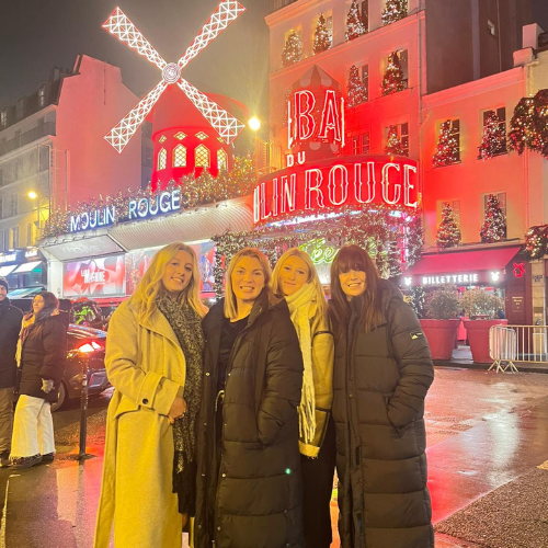 Four people posing at a rainy night market with bright neon signs and a windmill-lit building behind them