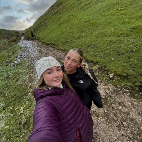 Two friends taking a selfie on a grassy hillside path under cloudy skies
