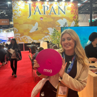 Woman at a Japan expo booth holding a pink MSG sign on a red carpet, smiling.