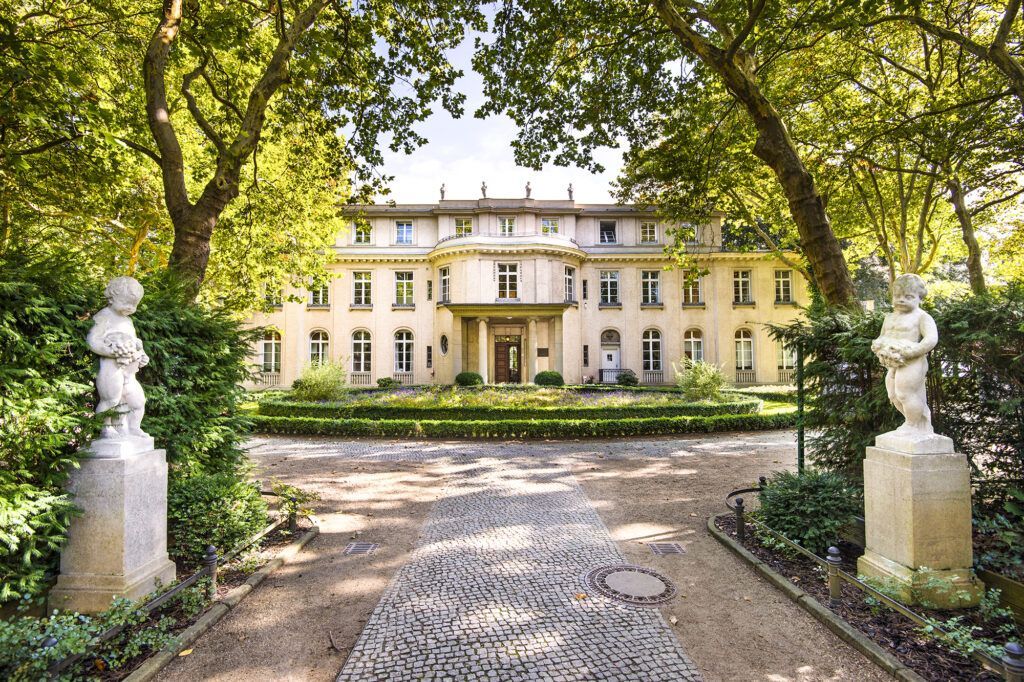 Historic cream mansion at end of a tree-lined garden path, framed by statues and arching trees.