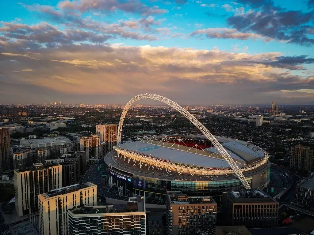 Aerial view of Wembley Stadium at sunset with its white arch and glowing lights over the city skyline