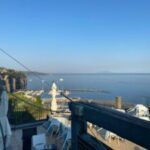 Coastal hillside view with blue sea, cliffs, and a white statue by a terrace railing
