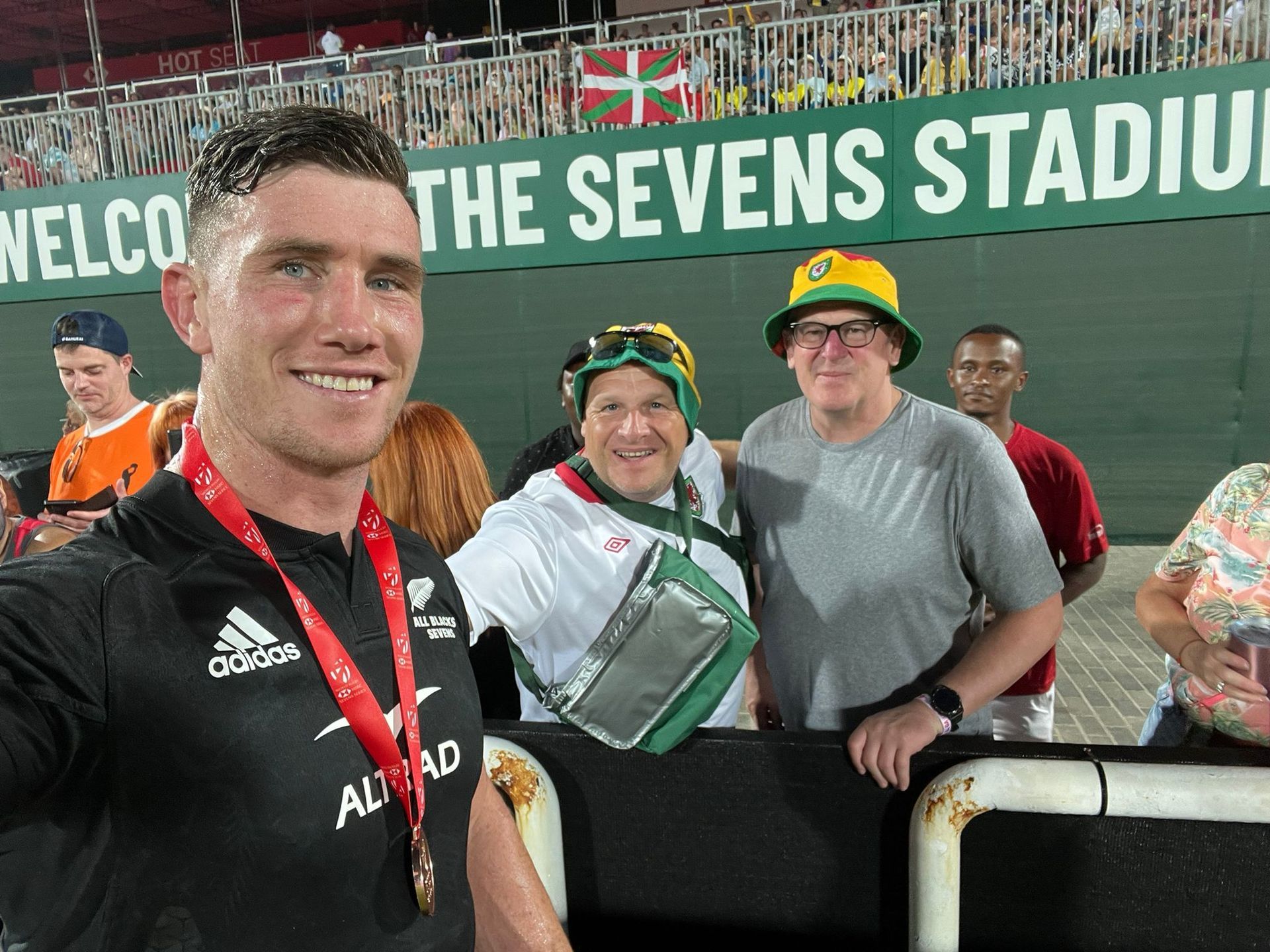 Smiling man in black jersey at The Sevens Stadium with fans behind him, holding a medal and gift bag