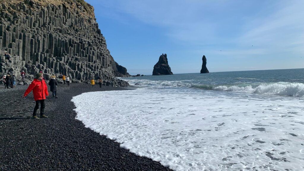 Person in a red jacket walking on a black pebble beach beside crashing waves and basalt cliffs.