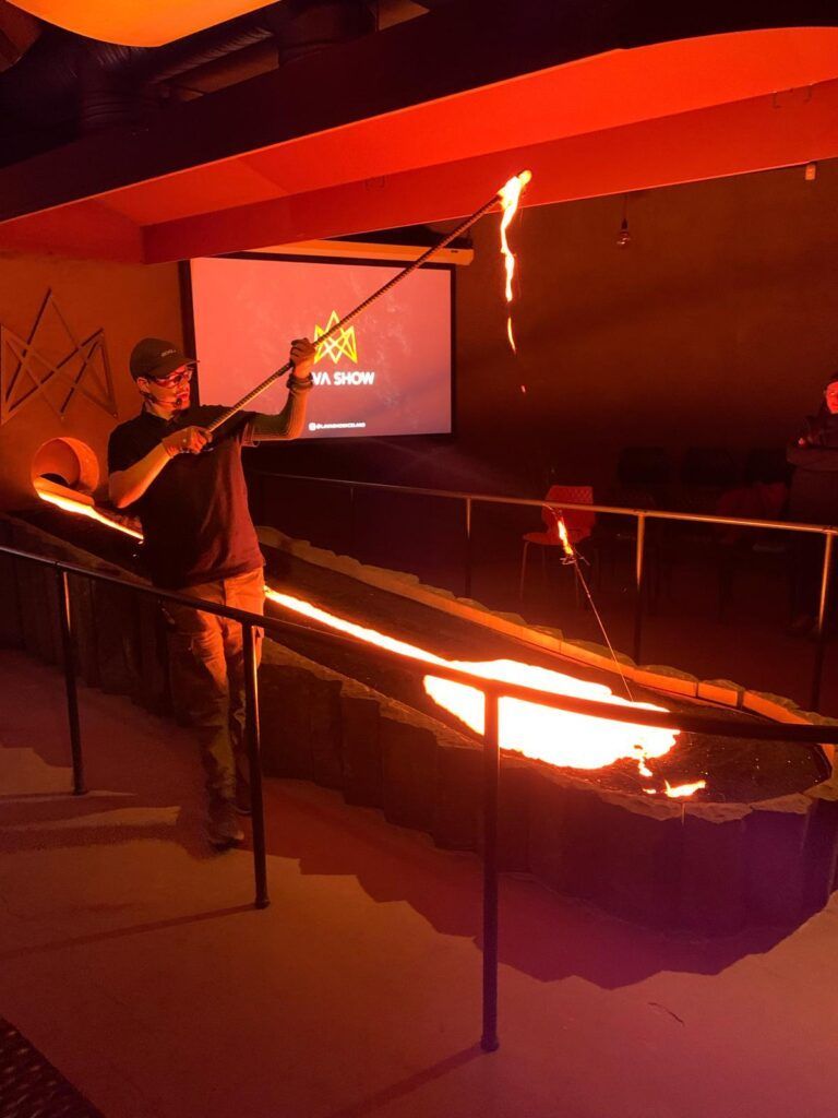 Person holding a torch over a long, glowing metal trough in a dim industrial workshop