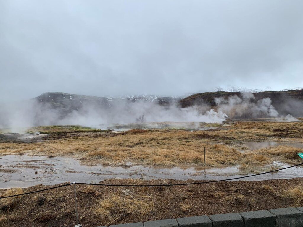 Steam rises from a barren geothermal field under a cloudy sky.