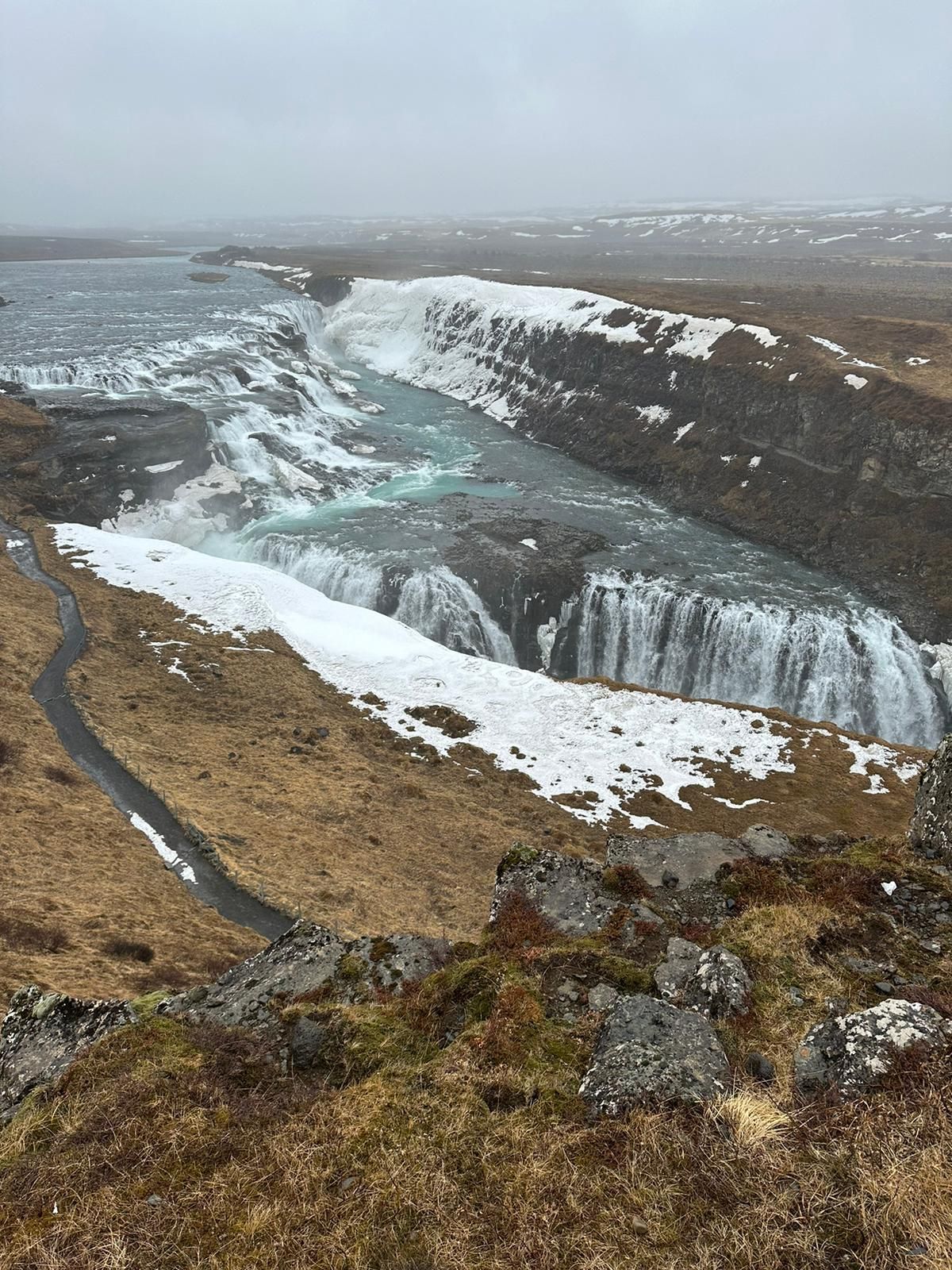 Icy waterfall and frozen river gorge in a snowy, rocky landscape under a gray sky