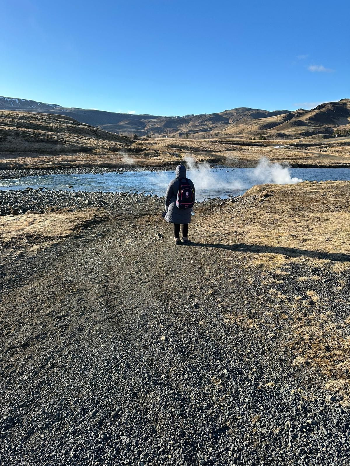 Person standing on a rocky shore by a steaming lake under a clear blue sky