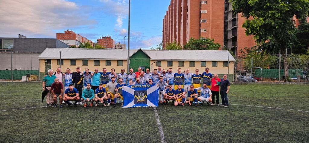 Group photo of a youth soccer team on a field with a banner, school buildings behind them.