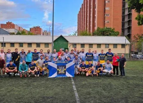 Group of rugby players and supporters posing on a field, holding a blue-and-white club banner.