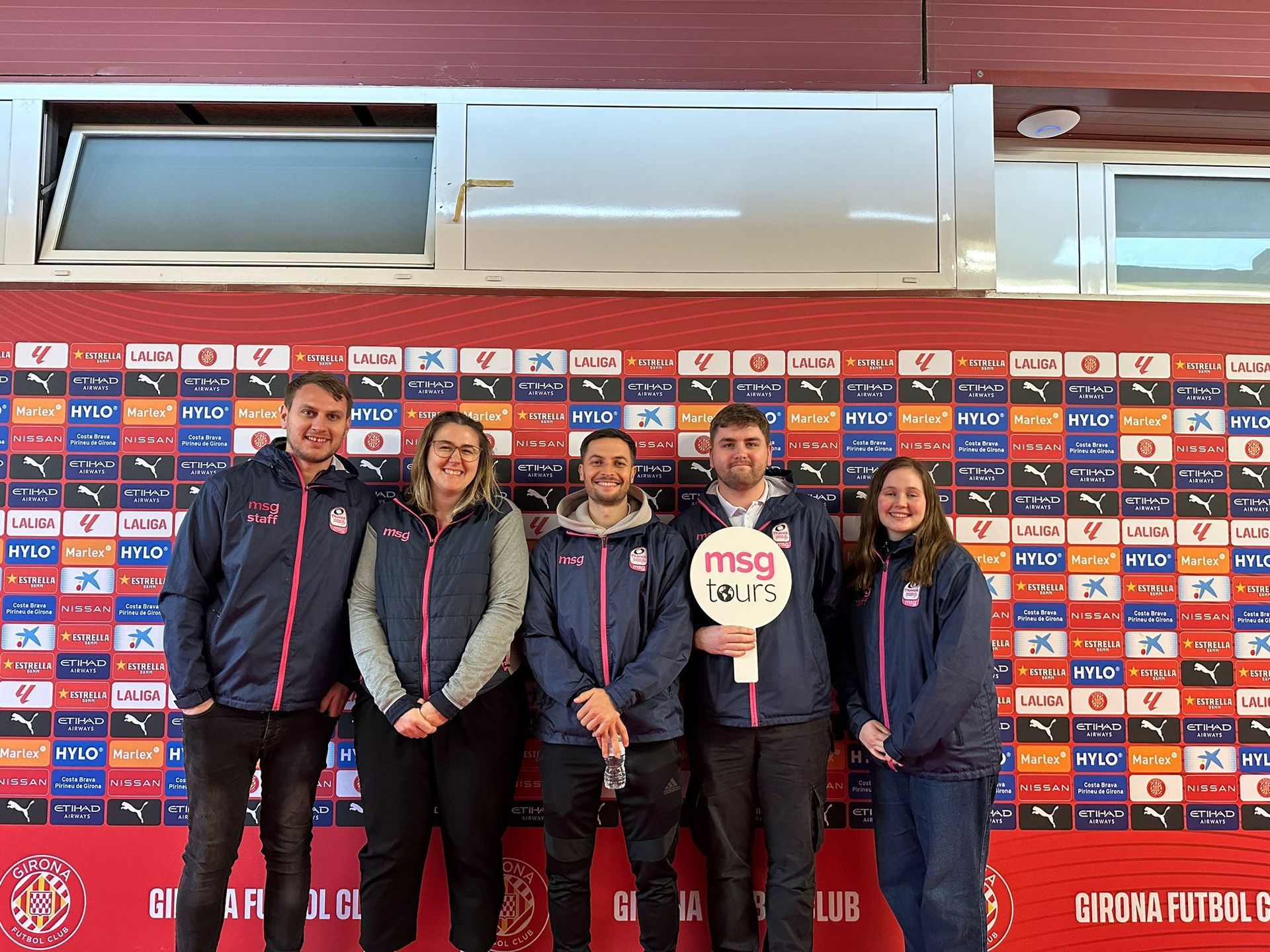 Five people posing in front of a sponsor backdrop at a sports venue, holding an 