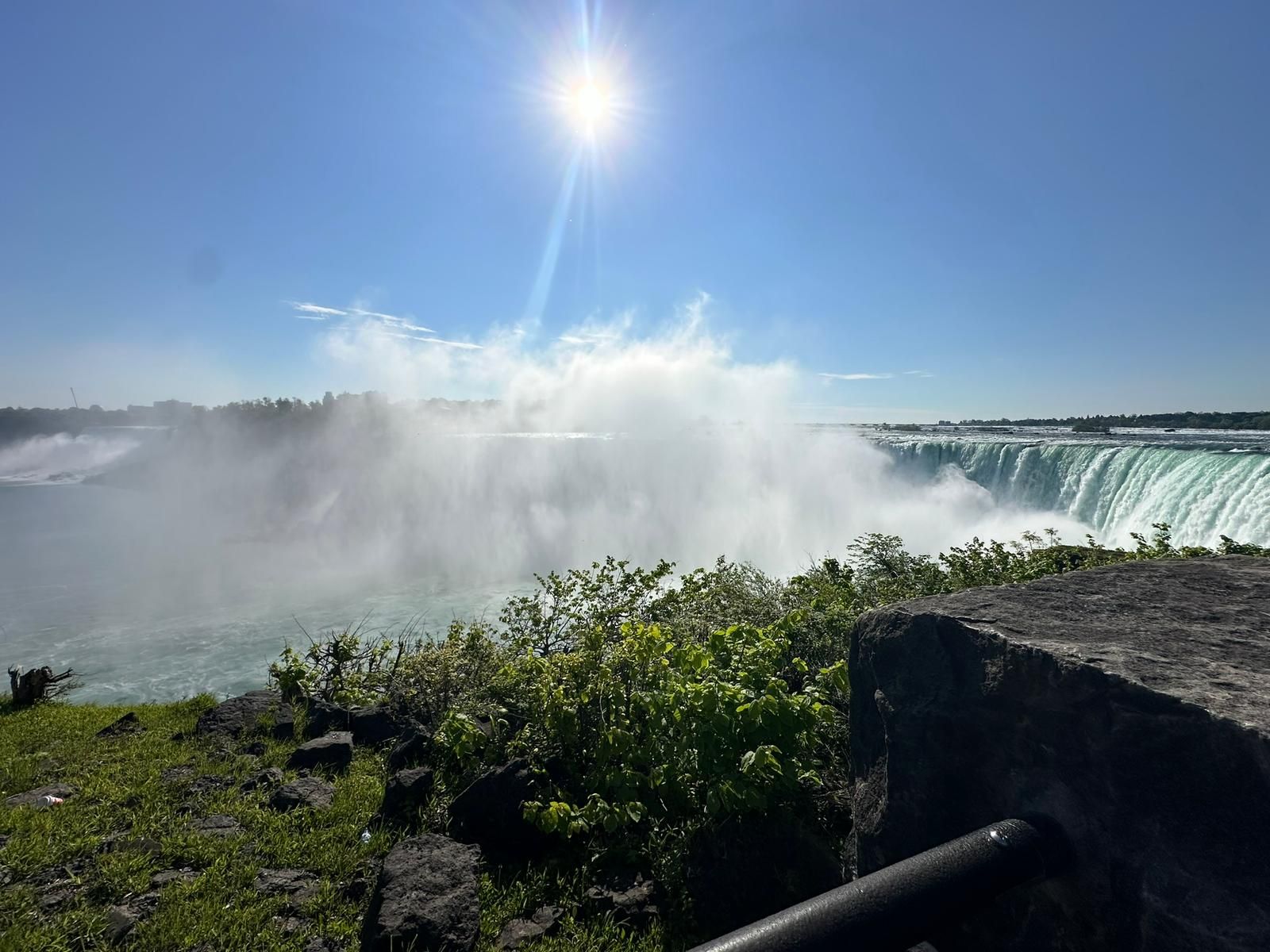 Sunlit waterfall with mist rising over a river, viewed from a rocky overlook with greenery