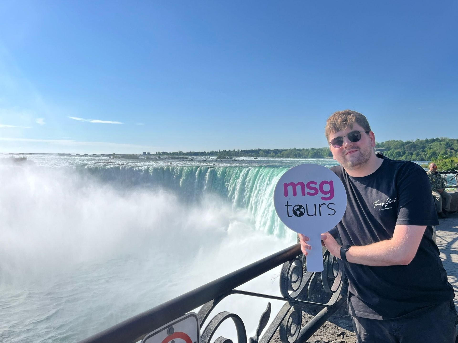 Person in sunglasses holding a sign by Niagara Falls on a sunny day, with mist rising from the water
