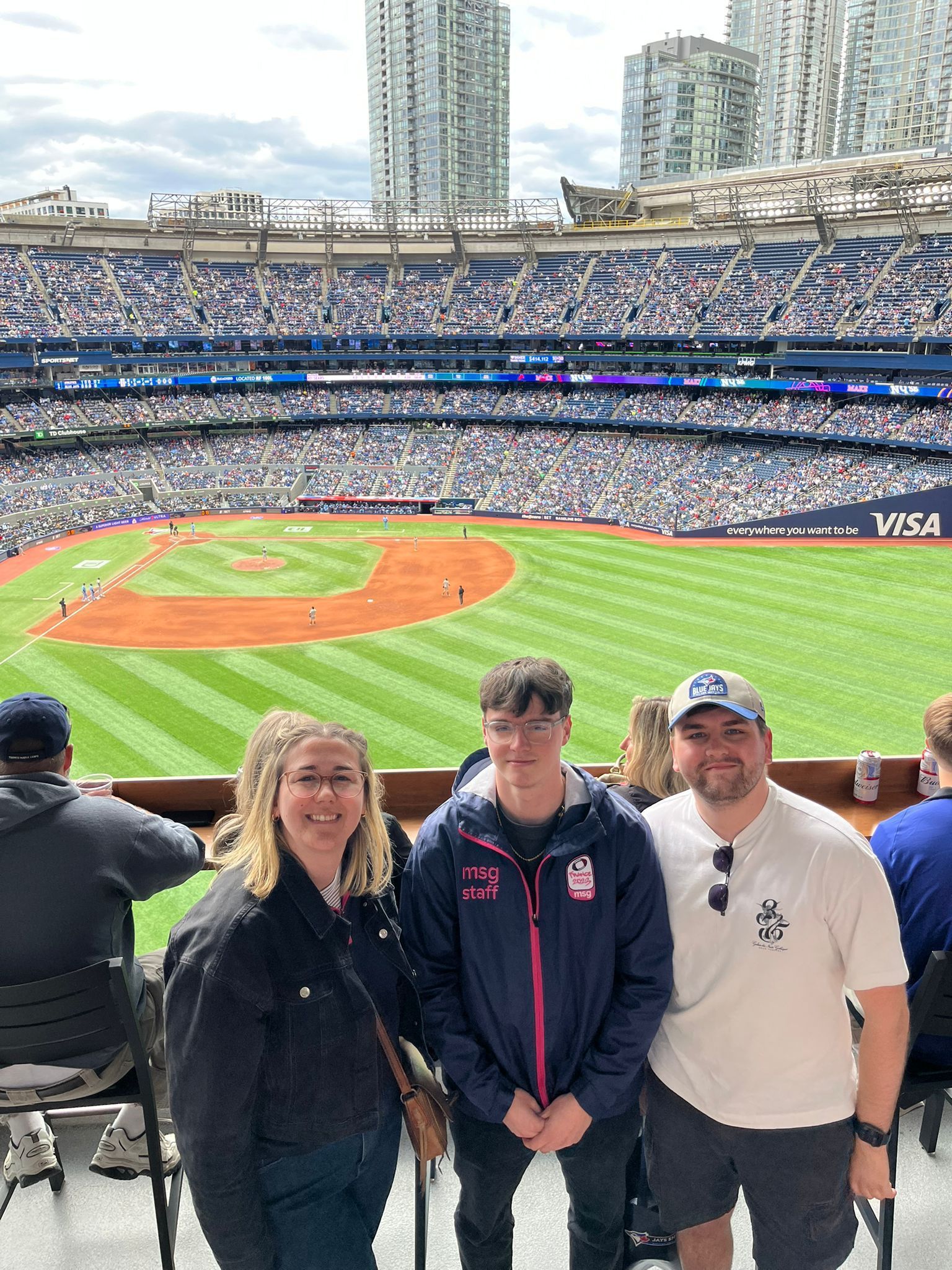 Three fans posing at a baseball stadium with the field and stands behind them