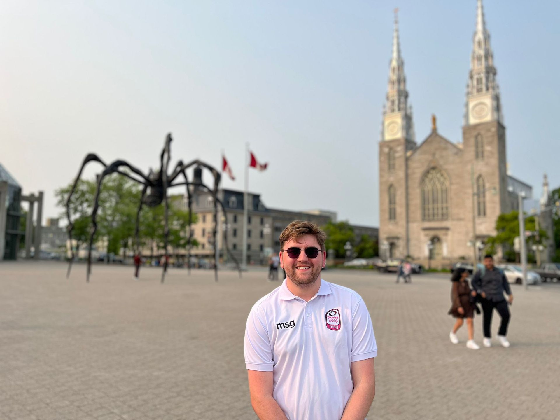 Man posing in sunglasses in a city square with a cathedral and spider sculpture in the background