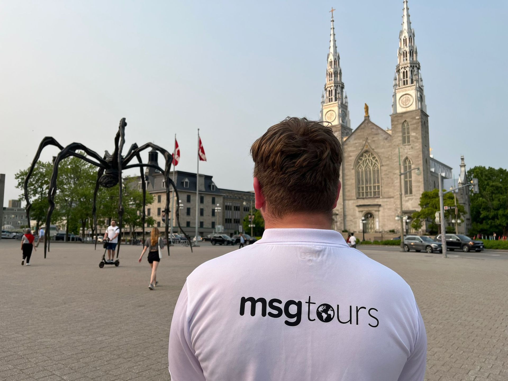 Person in white “msgtours” shirt facing a cathedral and giant spider sculpture in a city square
