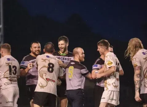 Soccer players in white jerseys celebrate together on a dark stadium field.