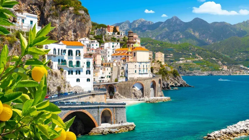 Colorful coastal village with white buildings, a stone bridge, turquoise sea, and mountains under a blue sky