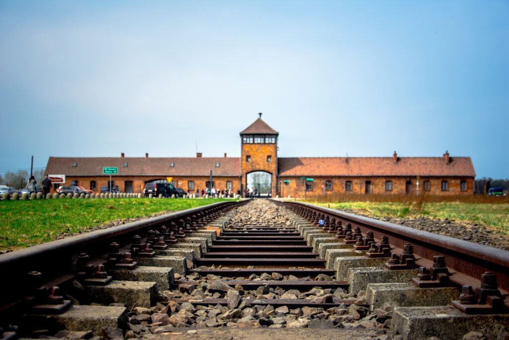 Railway tracks lead to a historic brick gate and long barracks under a cloudy sky