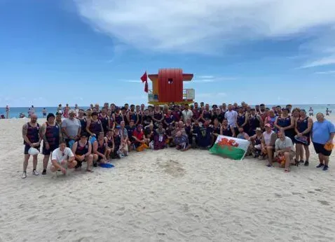 Large group posing on a sandy beach with a red lifeguard tower and blue sky in the background