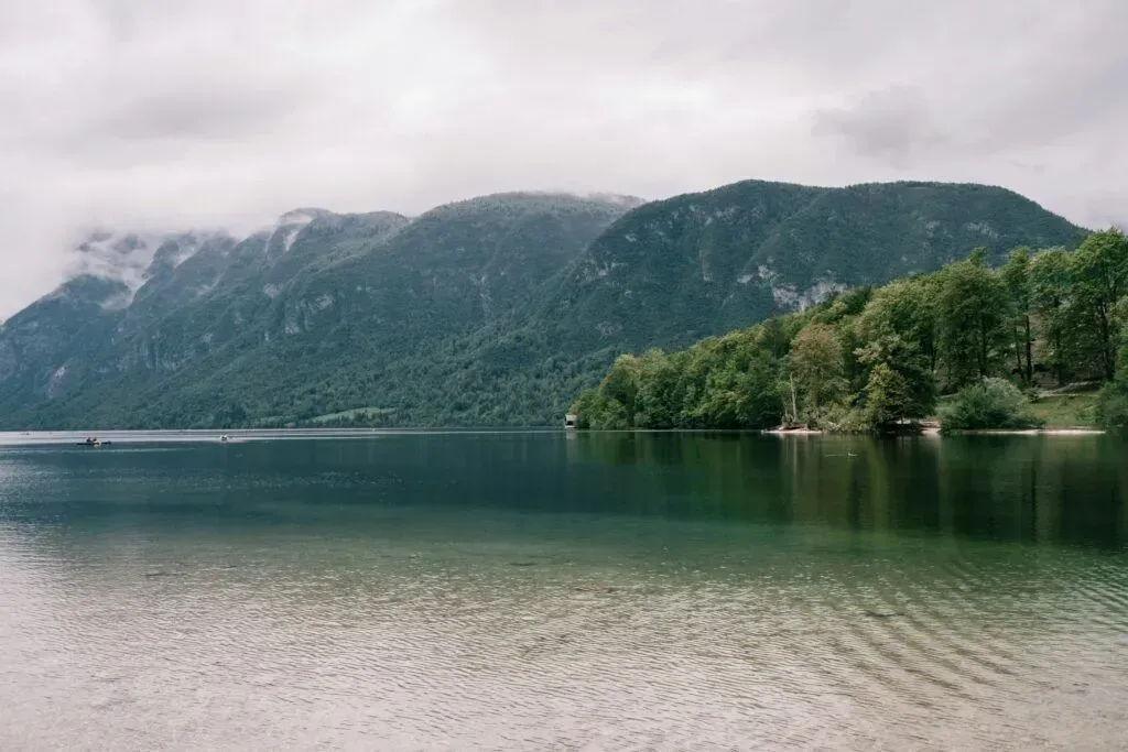 Calm lake with forested mountains under cloudy sky and a sandy shore in the foreground
