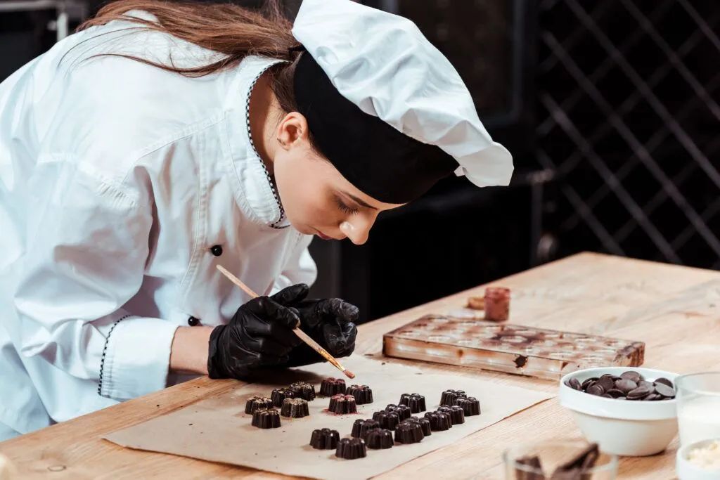 Chef in white uniform and hat arranging chocolates on a wooden board in a kitchen.