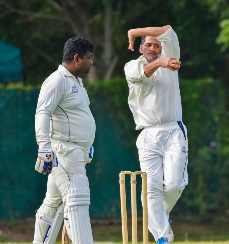 Two cricket players in white uniforms on a field, one bowling near the stumps and the other watching.