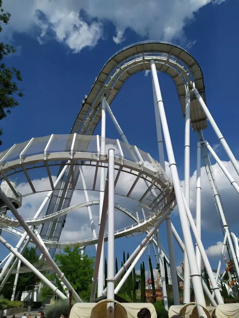 Steel roller coaster loops against a blue sky with clouds, surrounded by green trees.