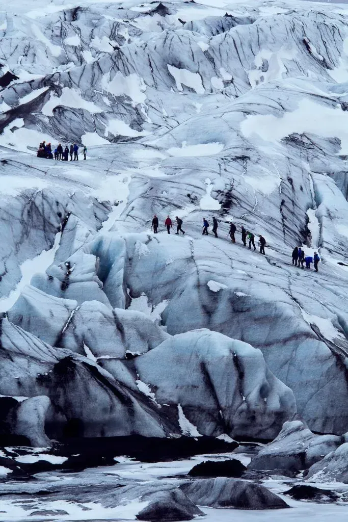 Small hikers crossing a snowy glacier with dark crevasses and rocky ice cliffs