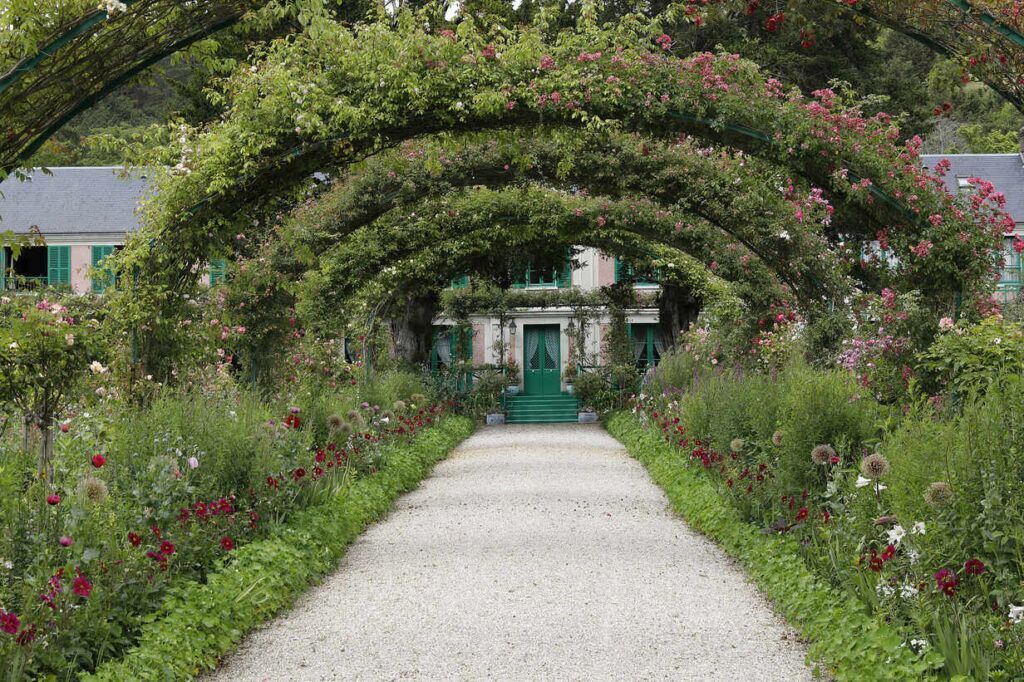 Garden walkway beneath a flower-covered arch leading to a greenhouse entrance