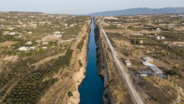 Aerial view of a long blue canal cutting through dry, sparsely built farmland with mountains in the distance