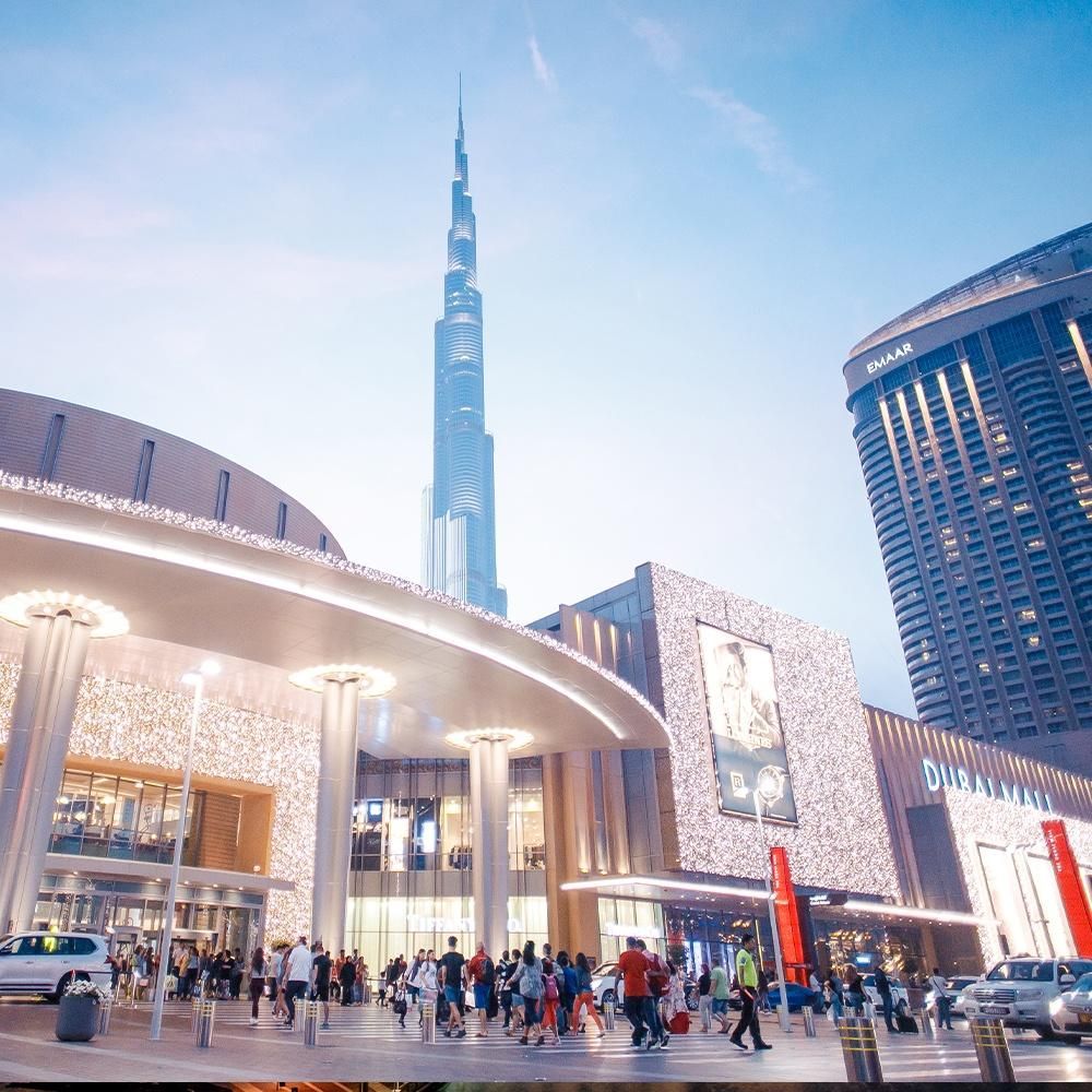 Crowded modern plaza with a curved mall entrance and Burj Khalifa rising in the background at dusk