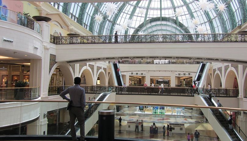 Interior of a large glass-roofed shopping mall with escalators and a few people walking below