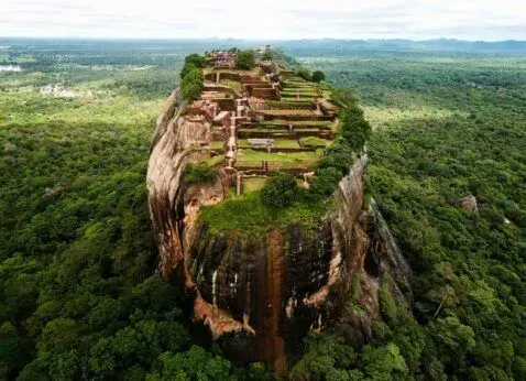 Ancient cliff-top ruins atop a steep rock, surrounded by dense green forest under a hazy sky